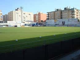 Vista exterior del campo de fútbol con gradas vacías y edificios urbanos al fondo