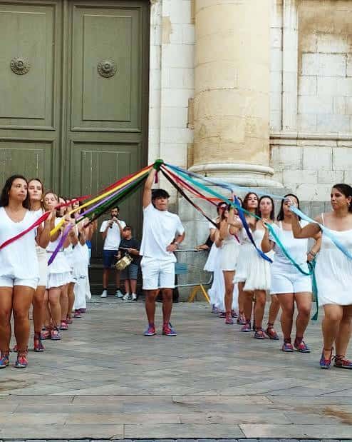 Grupo de bailarines con fajas de colores en una plaza frente a un edificio de piedra