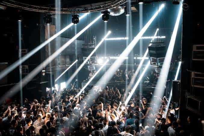 Escenario con haces de luz blanca cruzando la sala y multitud bailando