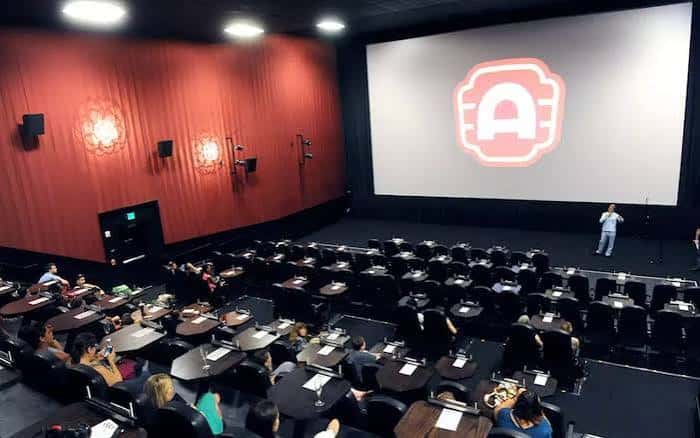 Sala de cine con butacas individuales en mesas y pantalla grande durante una proyección