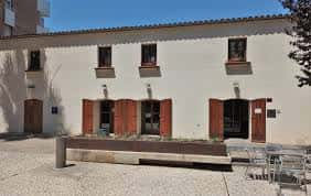 Vista exterior de la Plaça del Mestre Estalella con edificio de fachada blanca y ventanas con persianas marrones