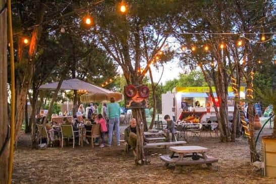 Área de comedor al aire libre con luces colgantes, camiones de comida y personas disfrutando del ambiente