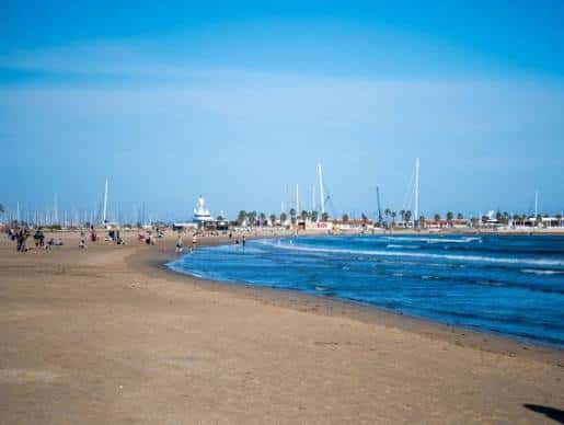 Vista panorámica de la playa con personas disfrutando y veleros al fondo