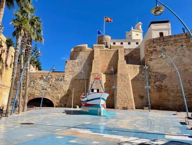 Plaza frente a la fortaleza con escultura de barco y banderas ondeando