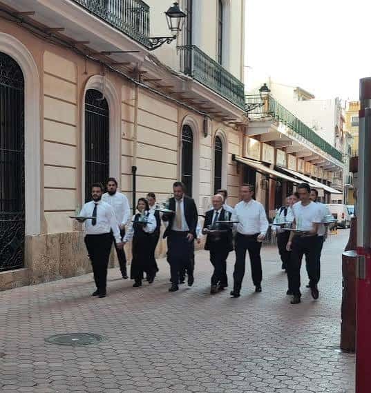 Grupo de personas caminando por la calle Carrer Escoles Pies durante un evento cultural o rodaje