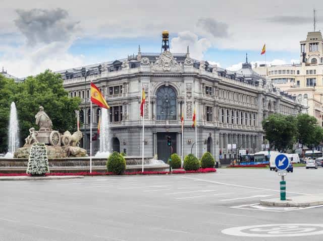 Vista frontal del edificio del Banco de España con fuente decorativa y múltiples banderas españolas en primer plano