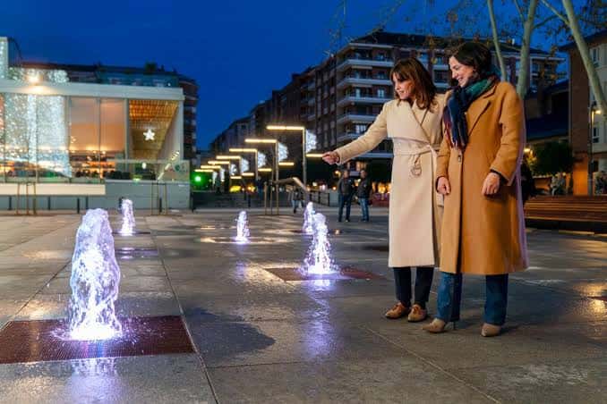Vista nocturna de la plaza con fuentes iluminadas y personas disfrutando del espacio