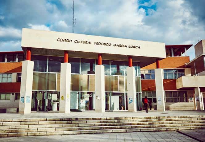 Vista frontal del Centro Cultural Federico García Lorca con su fachada de hormigón y columnas rojas bajo un cielo parcialmente nublado
