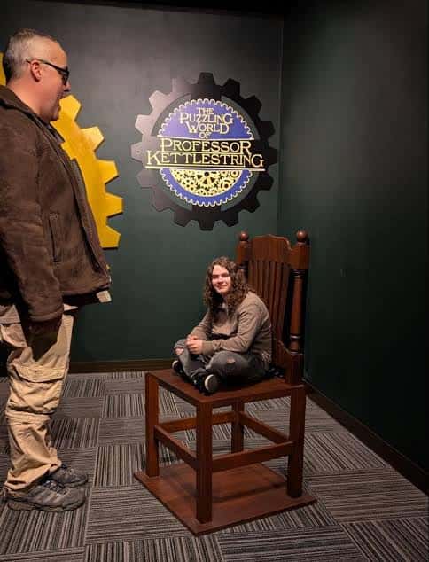 Hombre observando a una persona sentada en una silla pequeña frente al logo del museo