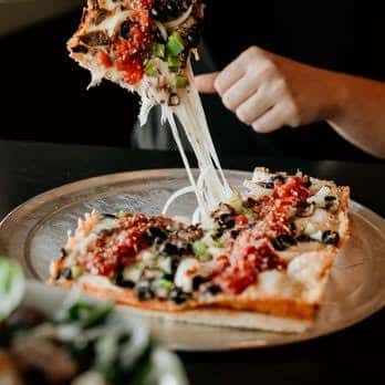 Rebanada de pizza cuadrada siendo levantada, mostrando queso derretido y toppings variados