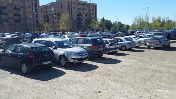 Aparcamiento exterior en tierra con múltiples coches bajo el sol y edificios residenciales al fondo