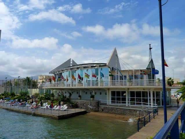 Vista exterior del club marítimo con terraza, banderas internacionales y zona de descanso junto al agua