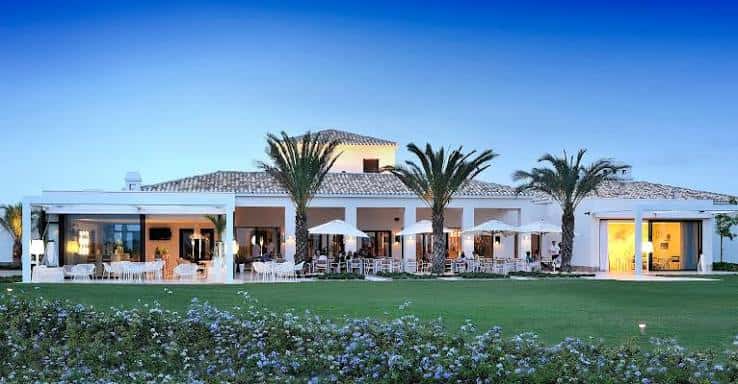 Terraza exterior iluminada del club con mesas y palmeras al atardecer