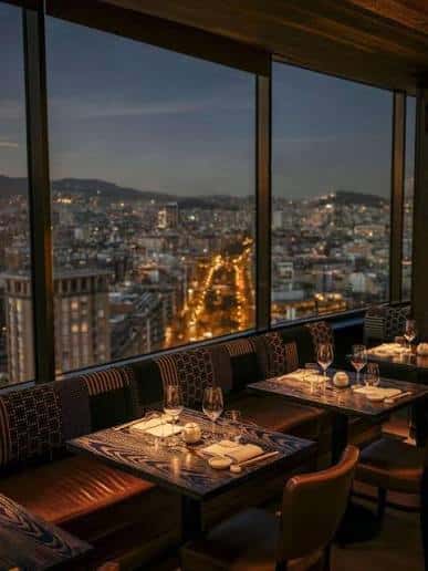 Interior elegante del restaurante del hotel con mesas servidas y vista nocturna de Barcelona desde grandes ventanales