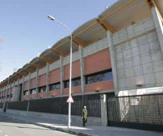 Fachada lateral del polideportivo bajo cielo despejado, con valla metálica y farola