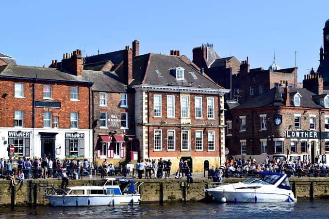 Fachadas de pubs y restaurantes en King's Staith con barcos amarrados y gente paseando