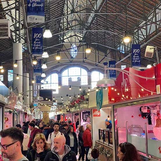 Interior del mercado con estructura metálica, luces colgantes y puestos comerciales decorados