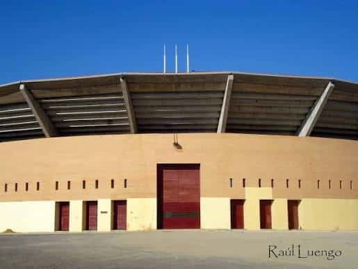 Fachada frontal de la plaza de toros bajo cielo azul, destacando sus puertas rojas y diseño arquitectónico