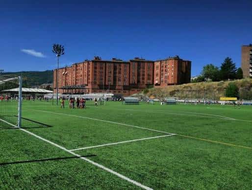 Vista general del campo de fútbol con jugadores en actividad y edificios residenciales al fondo