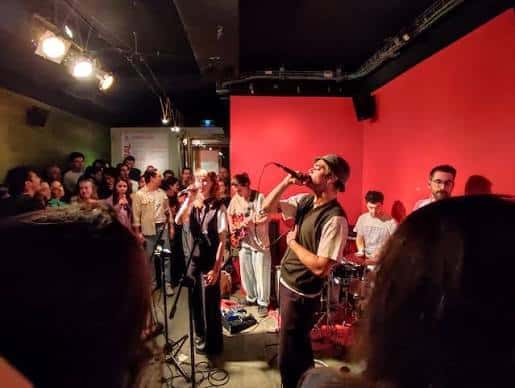Banda actuando en vivo frente a una pared roja y público de pie