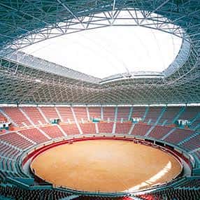 Interior del coso con tendido de arena y graderío rojo bajo estructura metálica
