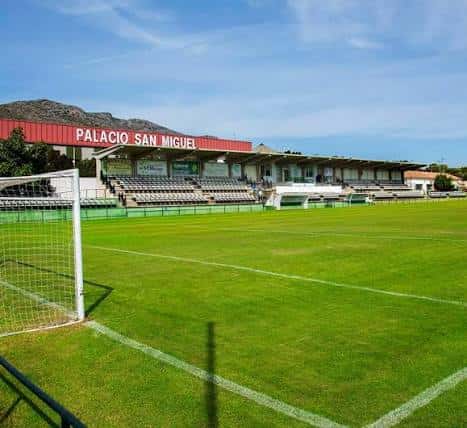 Campo de fútbol con césped verde y graderío bajo un cielo azul despejado