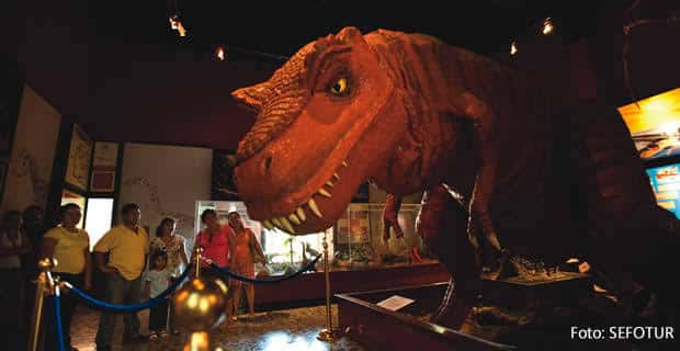 Exposición interior con réplica de T-Rex y visitantes observando la exhibición