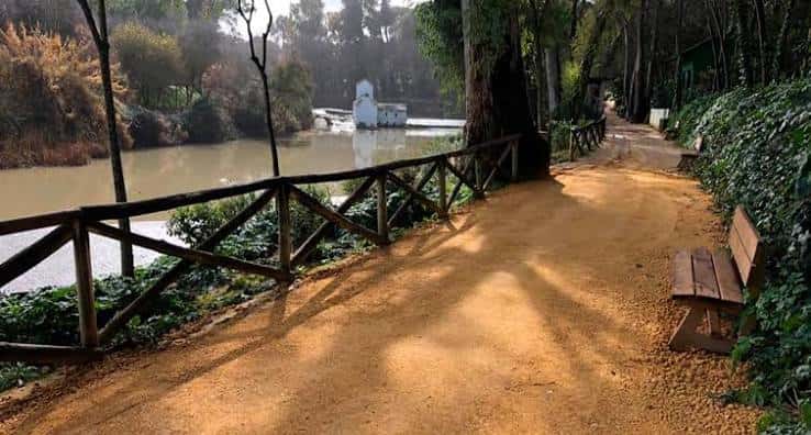Sendero de tierra con barandilla de madera bordeando el río y un banco de descanso
