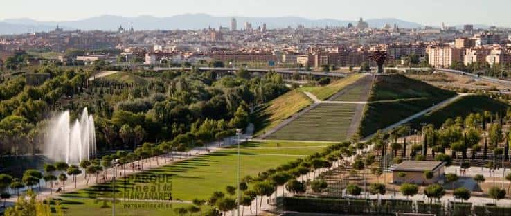 Vista aérea del parque con fuentes activas, senderos arbolados y la ciudad de fondo