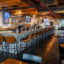 Área principal del bar con diseño industrial, vigas expuestas y pantallas de TV