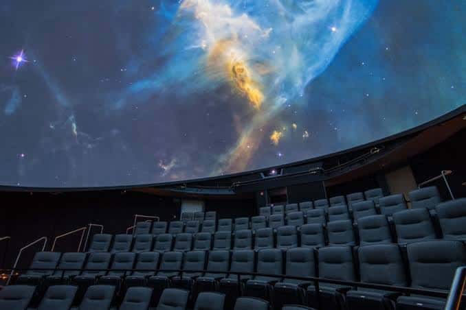 Interior del planetario con pantalla abovedada mostrando nebulosa estelar y butacas en gradas