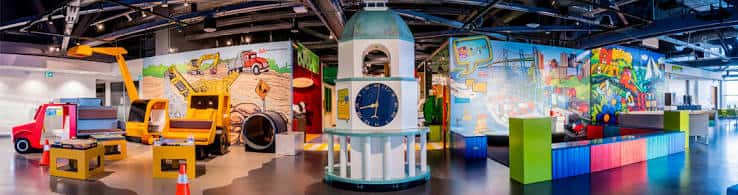 Zona infantil con réplicas de maquinaria amarilla, torre de reloj decorativa y murales temáticos