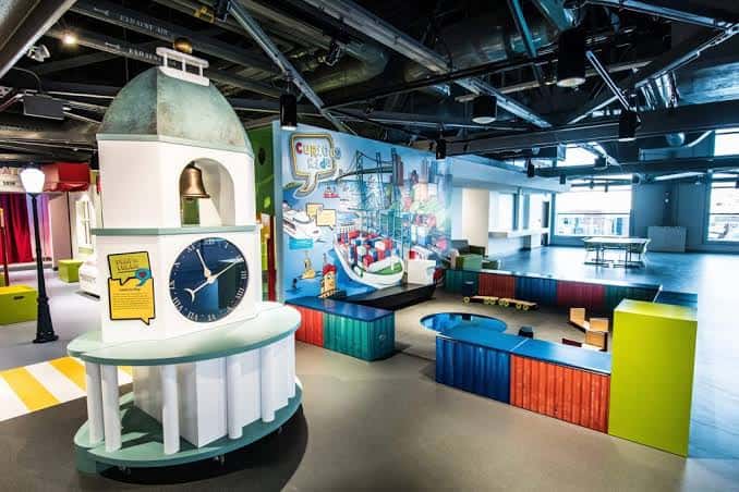 Área educativa 'Curious Kids' con torre de campanas, mesas de juego y mural de puerto marítimo