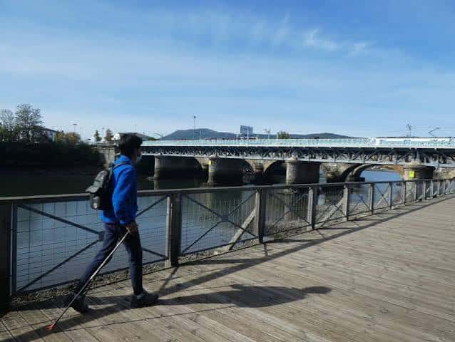 Persona caminando por la pasarela de madera junto al puente, con vista lateral del puente metálico