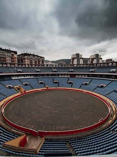 Plaza de toros desde las alturas con arena y graderío bajo nubes oscuras