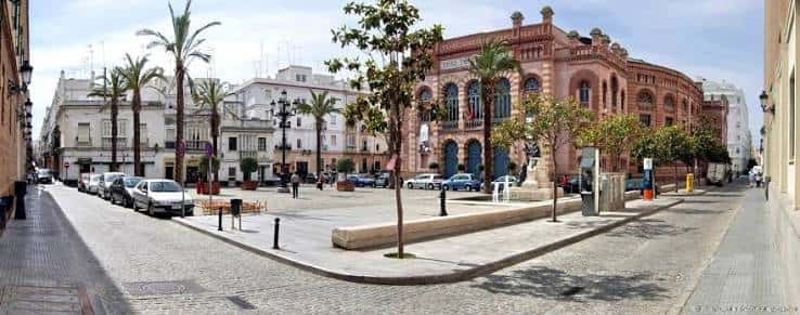 Vista lateral de la plaza con coches aparcados, árboles jóvenes y fachada del teatro