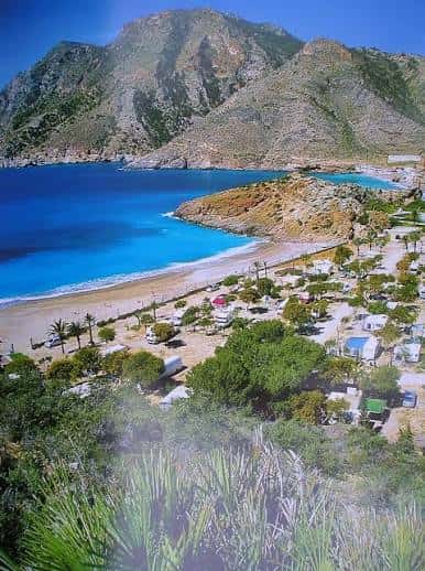 Panorámica del camping con playa frente al mar azul y montañas de fondo