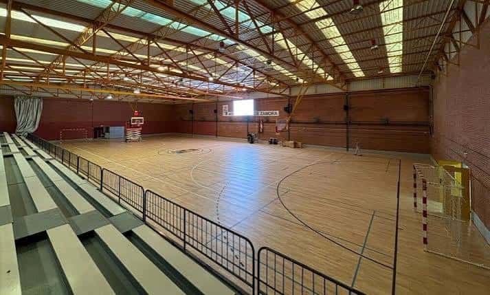 Interior del pabellón con pista de madera, gradas y estructura metálica visible en el techo