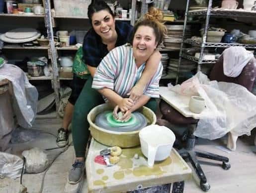 Dos mujeres sonriendo mientras trabajan en un torno de cerámica dentro del taller