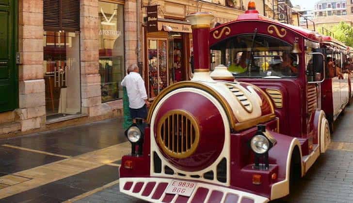 Vista lateral del tren turístico rojo circulando por una calle peatonal con tiendas