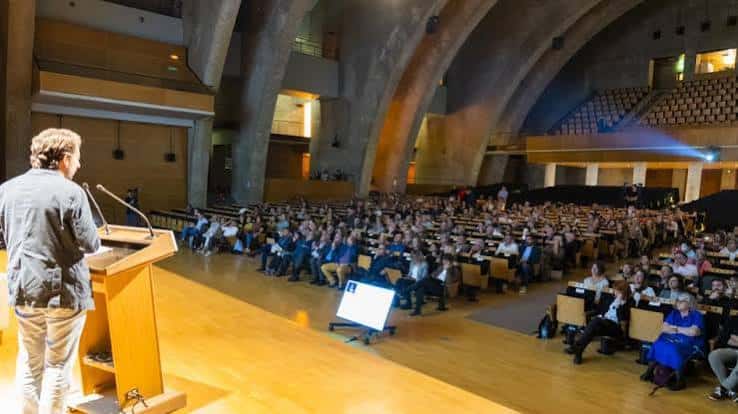 Interior del auditorio durante una conferencia, con público sentado y orador en el escenario