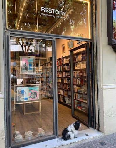 Vista exterior de la librería Restory con su escaparate iluminado y un perro sentado en la entrada