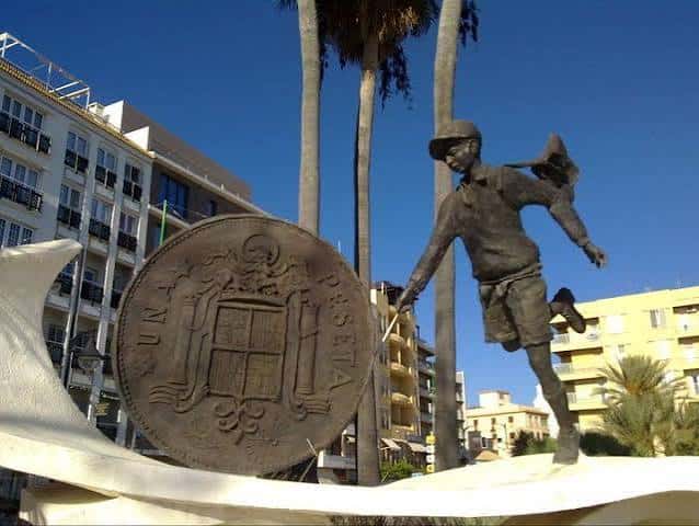 Plano medio de la escultura mostrando al niño sosteniendo una peseta frente a edificios residenciales