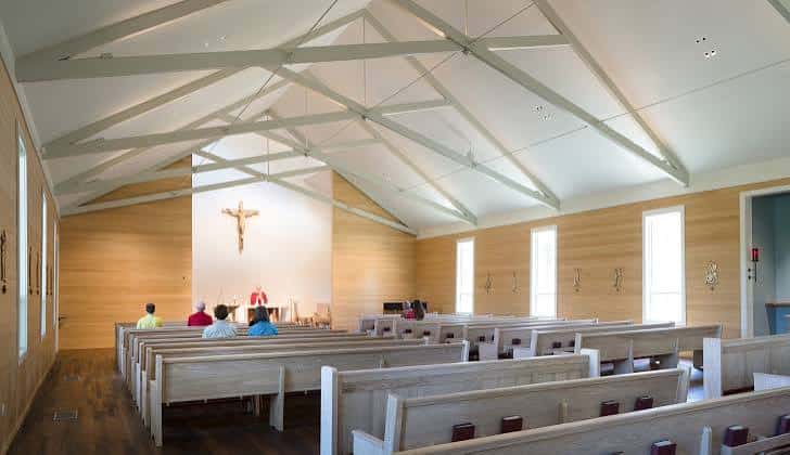 Interior moderno de capilla con techos altos, vigas blancas y bancos de madera clara