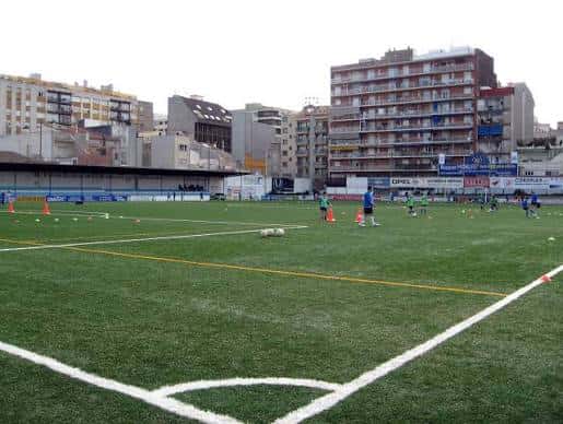 Campo de fútbol con jugadores entrenando, gradas y edificios residenciales al fondo