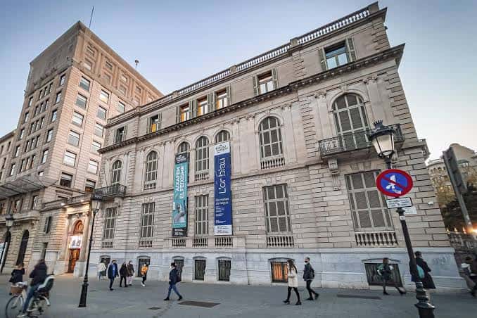 Fachada del Palau Robert con carteles de exposiciones y peatones en la acera