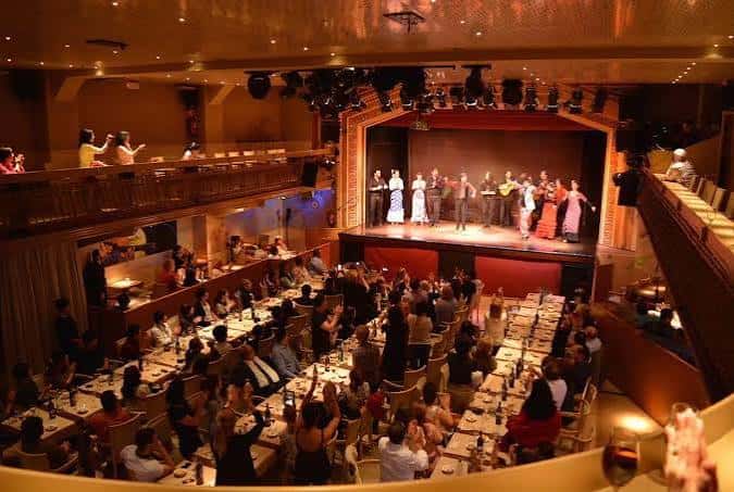 Vista general del interior del teatro durante un evento con público en mesas y artistas en el escenario