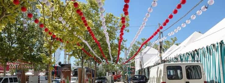 Pasillo decorado con farolillos rojos y blancos entre árboles y casetas