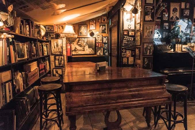 Escenario íntimo del café con piano antiguo, taburetes y paredes adornadas con fotografías y recuerdos musicales