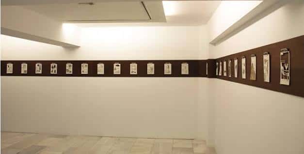 Interior de sala de exposiciones con obras colgadas en pared oscura sobre fondo blanco